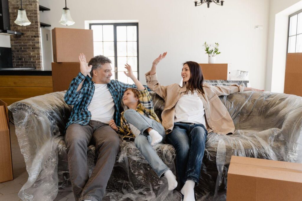 A father, mother, and young son high-five each other while sitting on a plastic-wrapped sofa surrounded by moving boxes in their new living room.